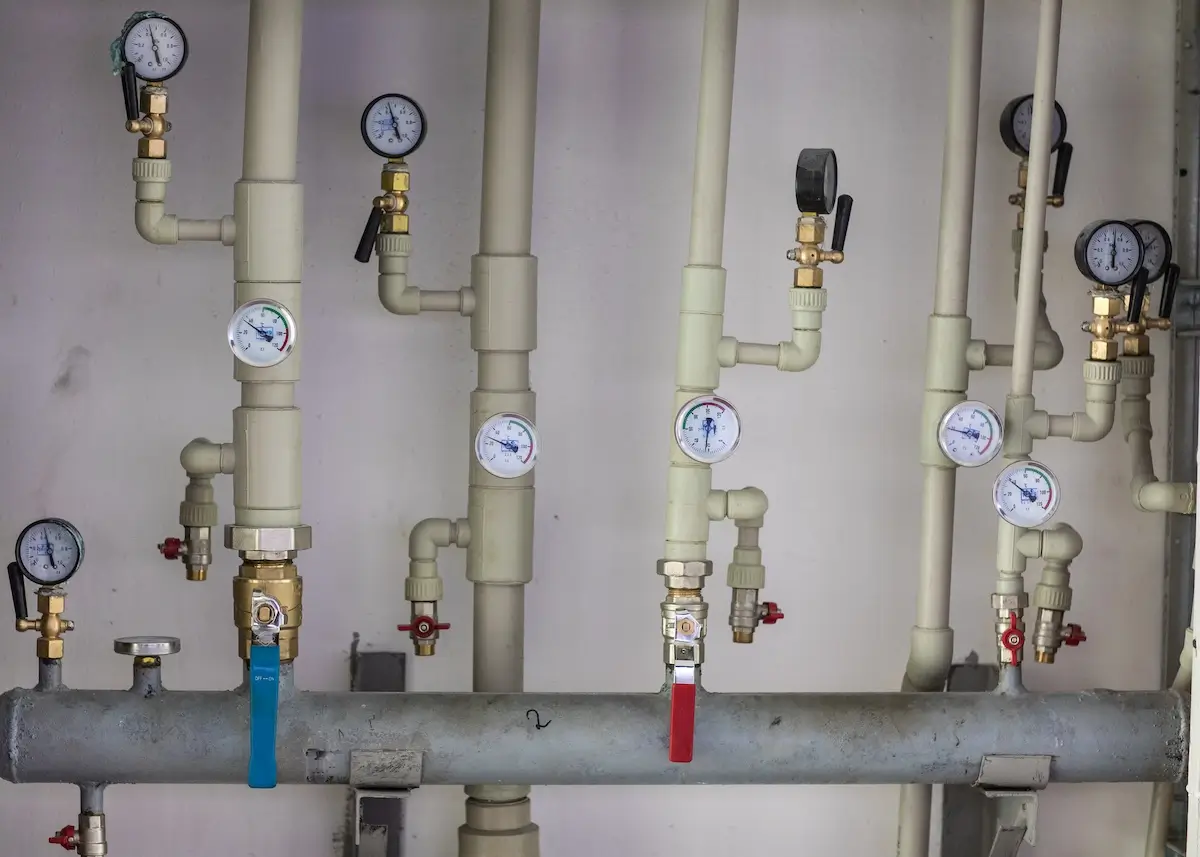 Several white pipes with pressure gauges, valves, and levers are connected to a horizontal metal manifold against a plain wall. The gauges display various pressure readings, and some valves have blue or red handles.