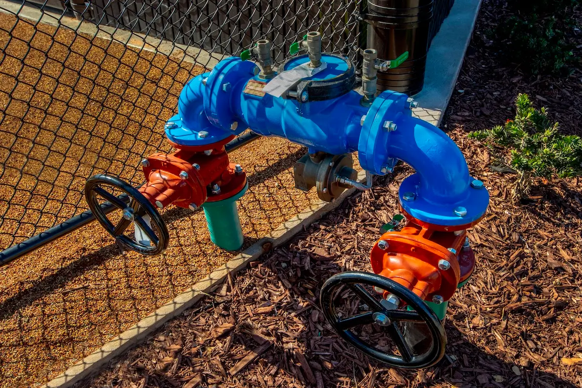 A blue water backflow preventer assembly with two large red valve handles and black wheel controls is installed on the ground next to a chain link fence, surrounded by mulch and gravel.