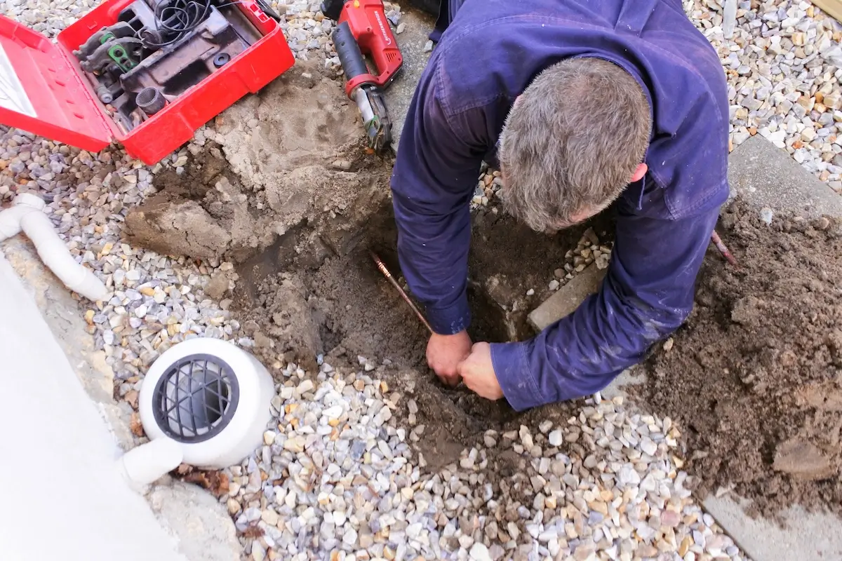 A person with gray hair wearing a blue shirt is kneeling on gravel, repairing pipes in a shallow trench. Tools and a red toolbox are nearby.