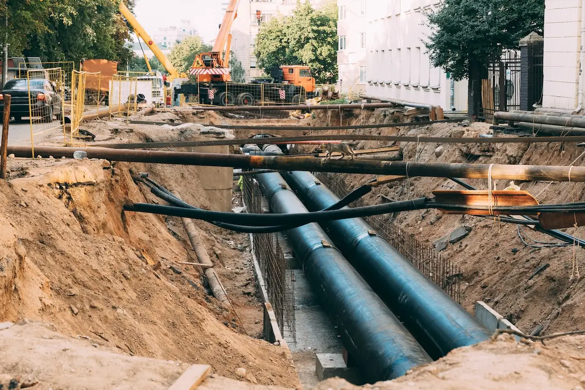 Large pipes being installed underground at a construction site in a city, with cranes, construction materials, and workers visible in the background. Dirt is piled along the edges of the trench.
