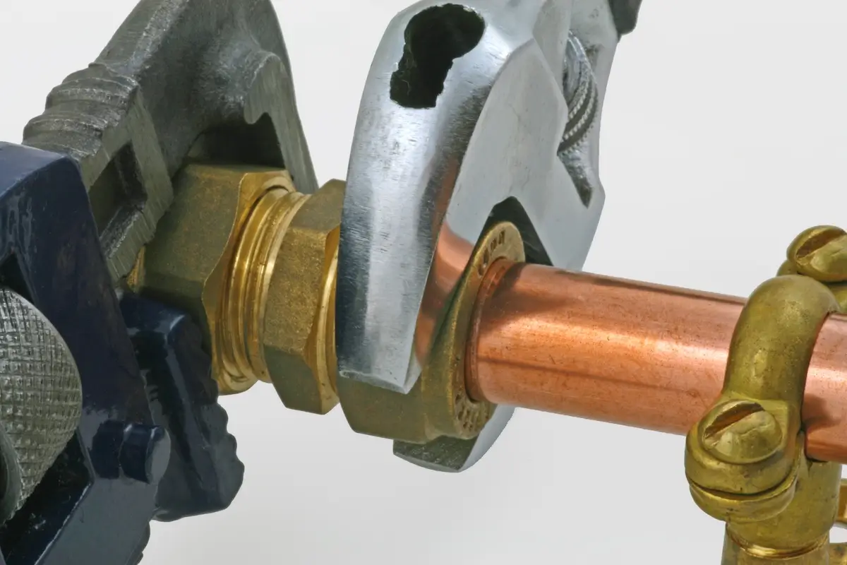 Close-up of two wrenches tightening a brass fitting onto a copper pipe, with an additional brass valve assembly attached to the pipe, set against a plain white background.