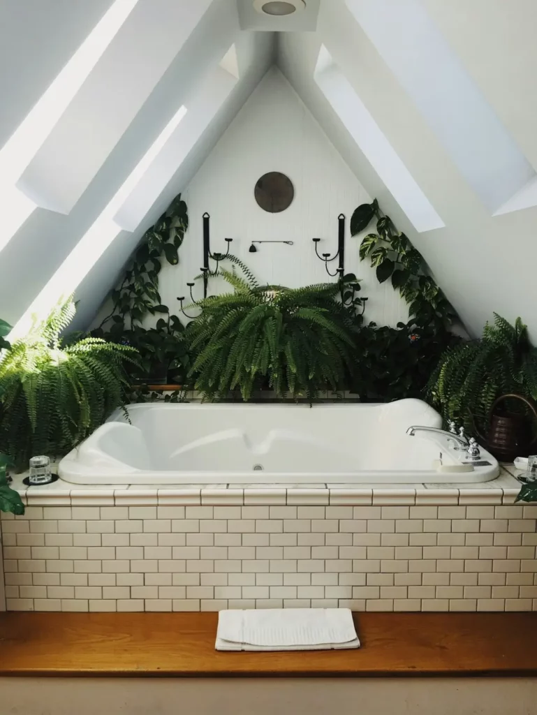 A white bathtub with tiled surround sits under a sloped ceiling with skylights, surrounded by lush green plants and ferns, creating a serene, spa-like atmosphere. A wooden bench and a white bath mat are in front.