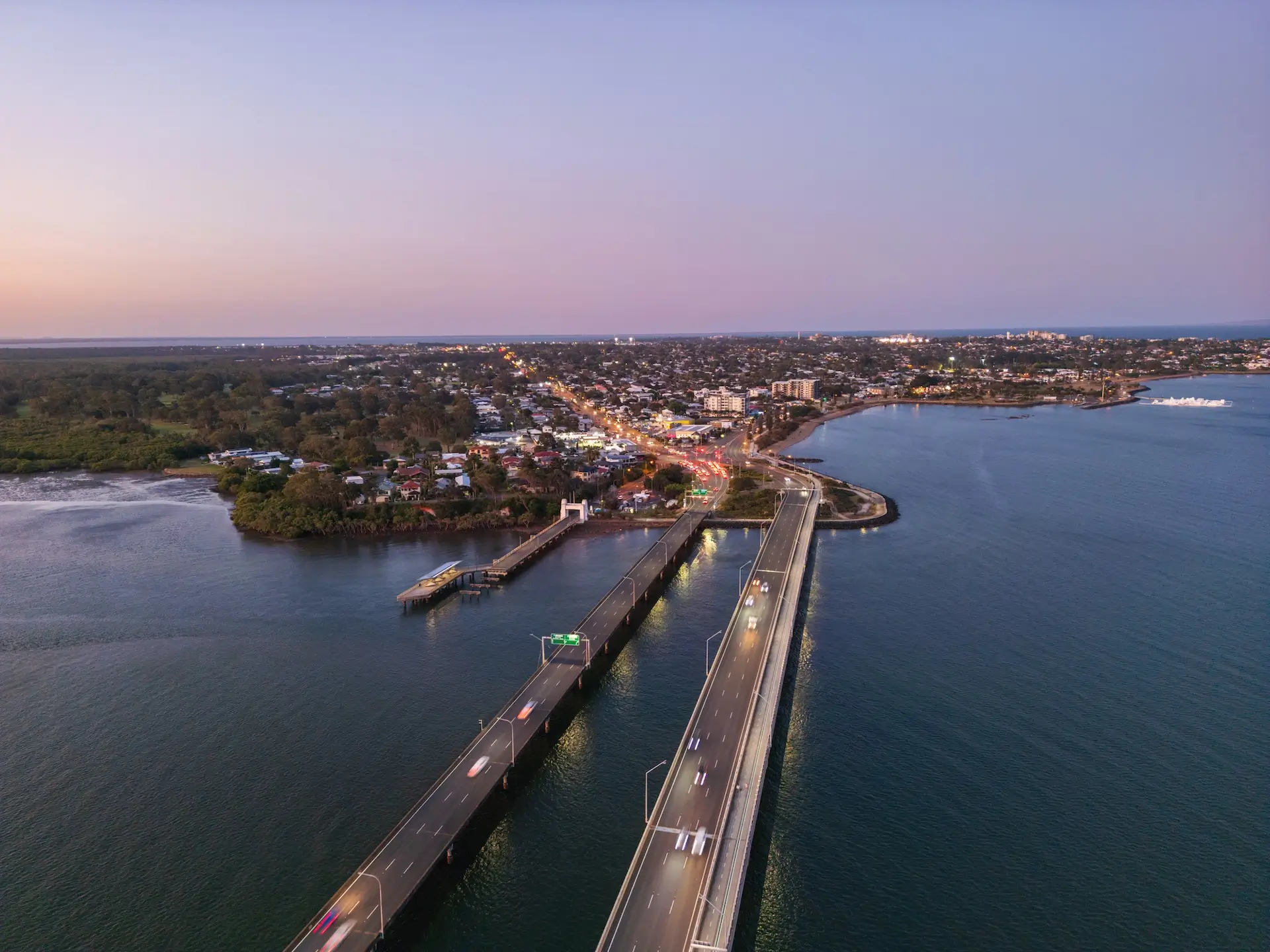 Aerial view of a curved coastal road and bridge over water at dusk, with cars driving, a town with scattered lights, and trees on the left and open sea on the right. The sky is clear and tinged with soft evening colors.