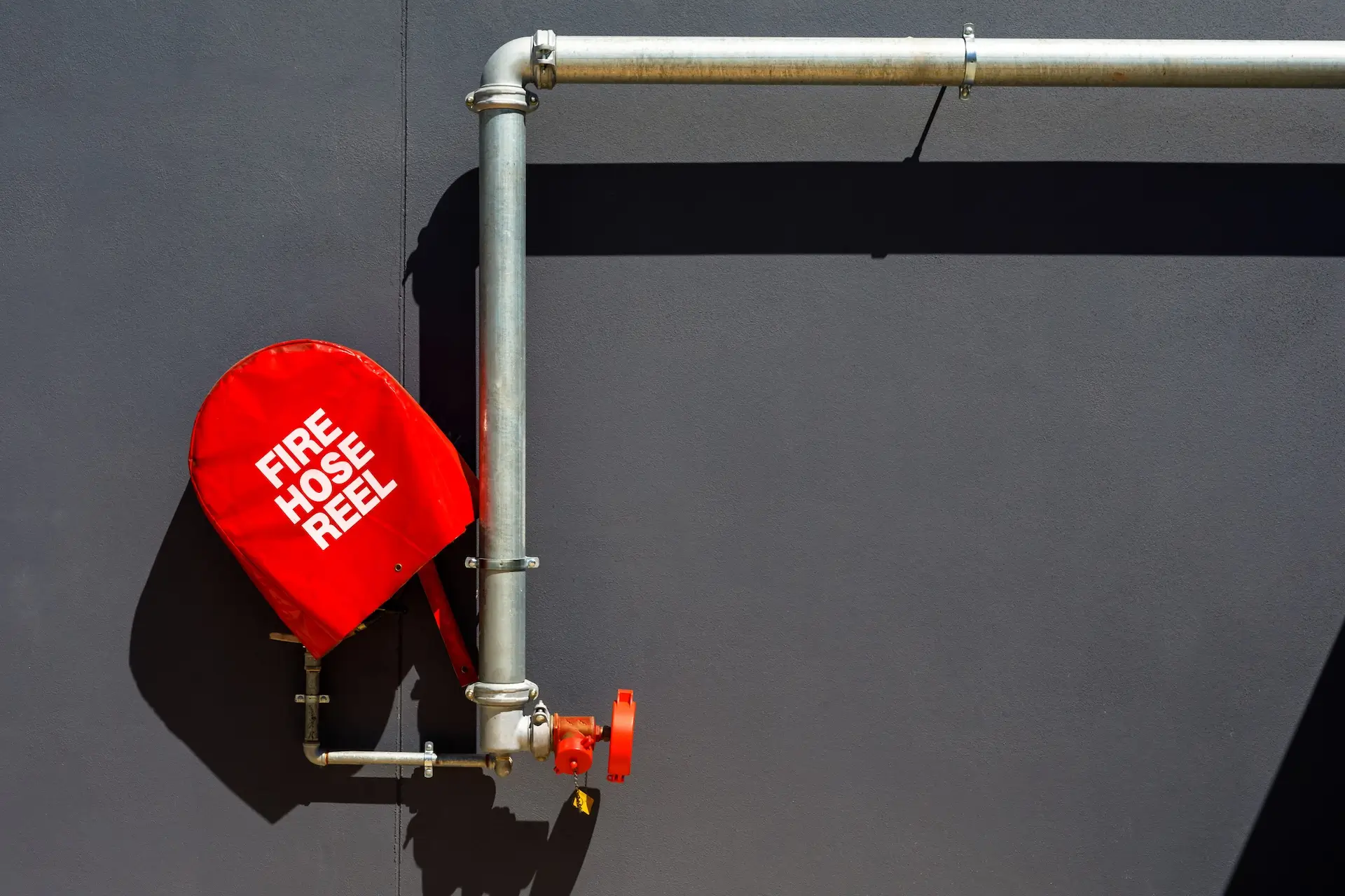 A red fire hose reel with white text is mounted on a grey wall, connected to horizontal and vertical metal pipes with a red valve handle.