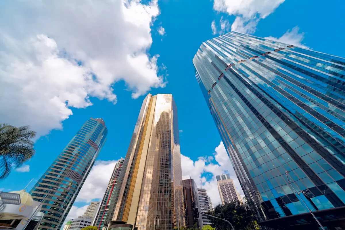 Tall modern skyscrapers with glass facades reflect sunlight against a bright blue sky with scattered clouds, viewed from a low angle in a cityscape.