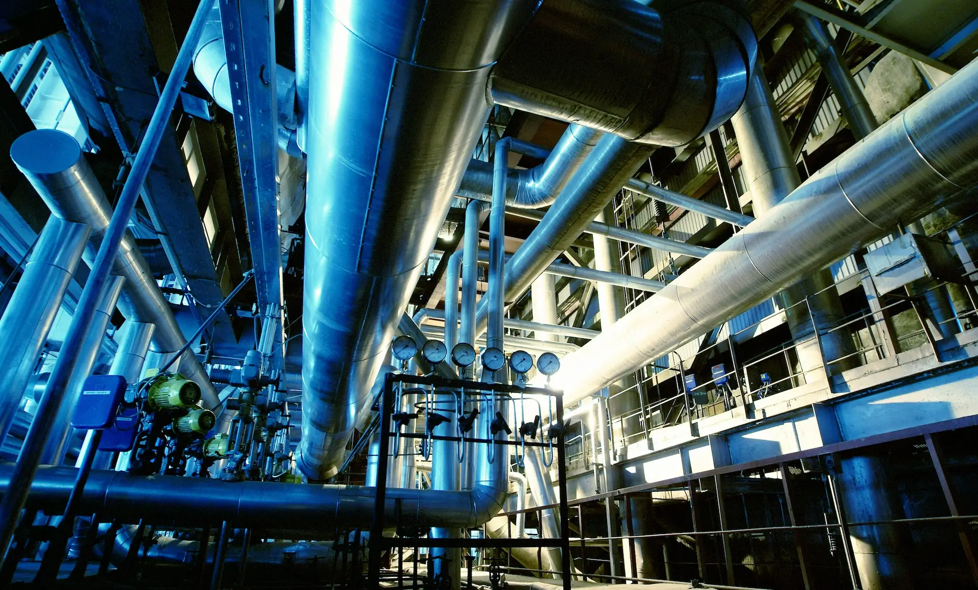 Complex network of large, shiny metal pipes and industrial gauges in a brightly lit processing plant or factory, with platforms and railings for maintenance access.