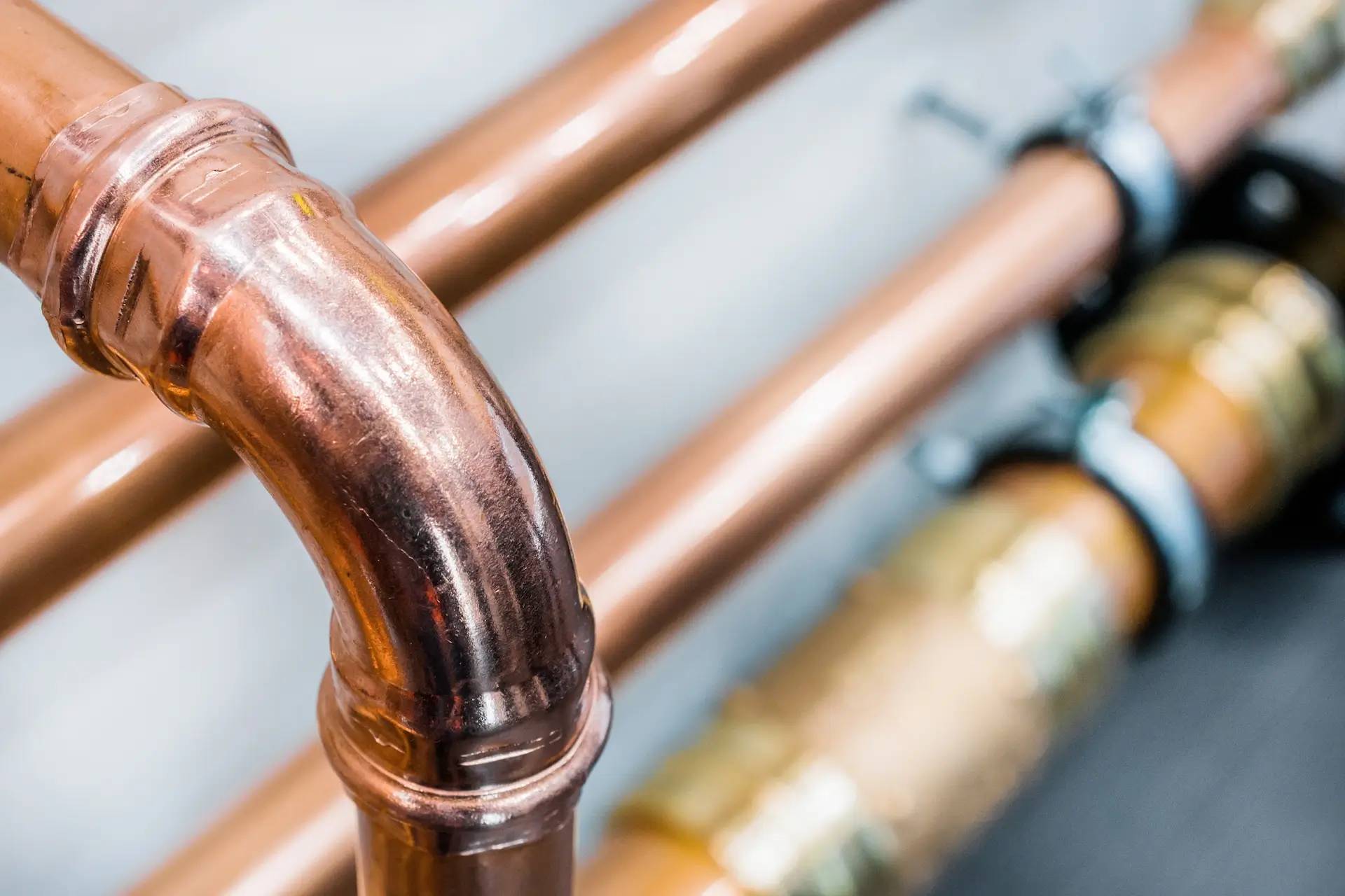 Close-up of shiny copper pipes and fittings, including a curved elbow joint, with additional pipes in the blurred background secured by metal brackets.