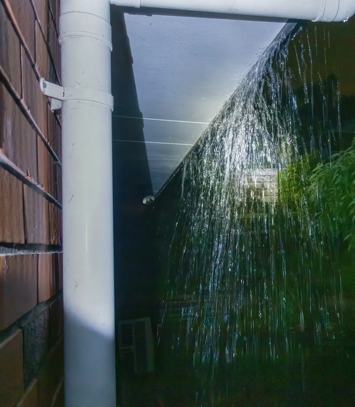 Water is leaking from the joint of a white PVC downpipe attached to a brick wall at night, creating a spray of water illuminated by light. A leafy tree is visible in the background.