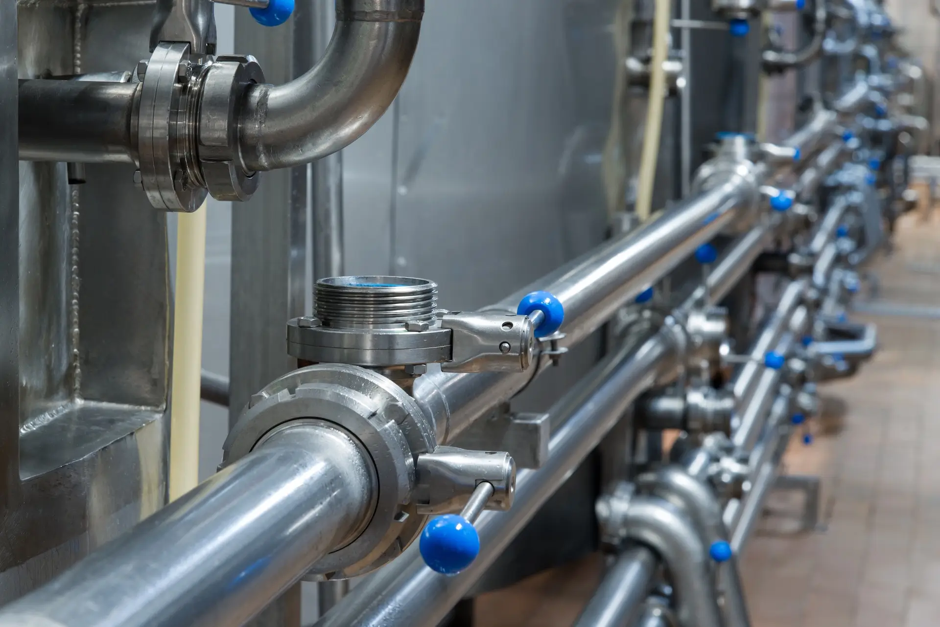 Close-up view of industrial stainless steel pipes and valves with blue handles in a factory setting, showing a clean and modern processing or manufacturing environment.